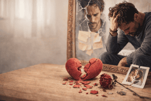 Mann sitzt nach einer Trennung traurig vor einem zerbrochenen Herz auf einem Tisch und denkt über eine Beziehung mit einem Narzissten nach